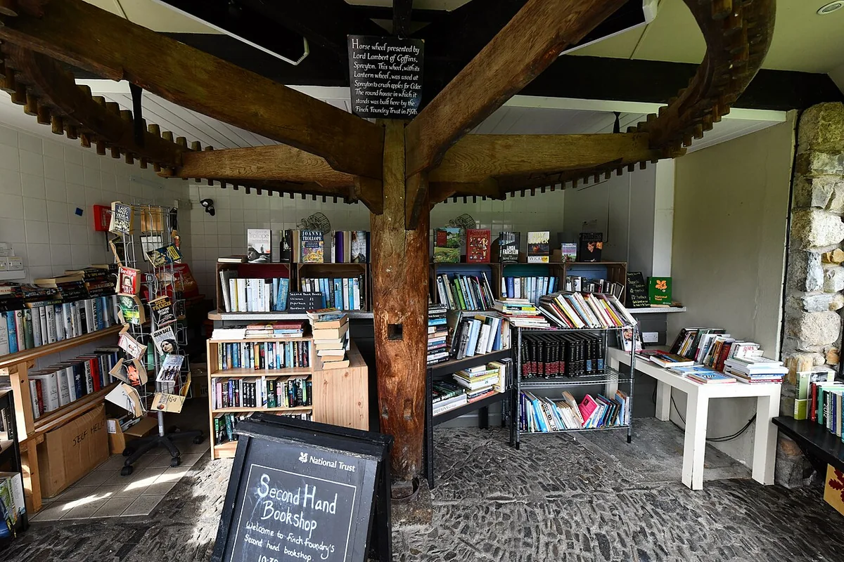 Shelves and tables of second-hand books under a large wooden horse wheel, with a National Trust 'Second Hand Bookshop' sign
