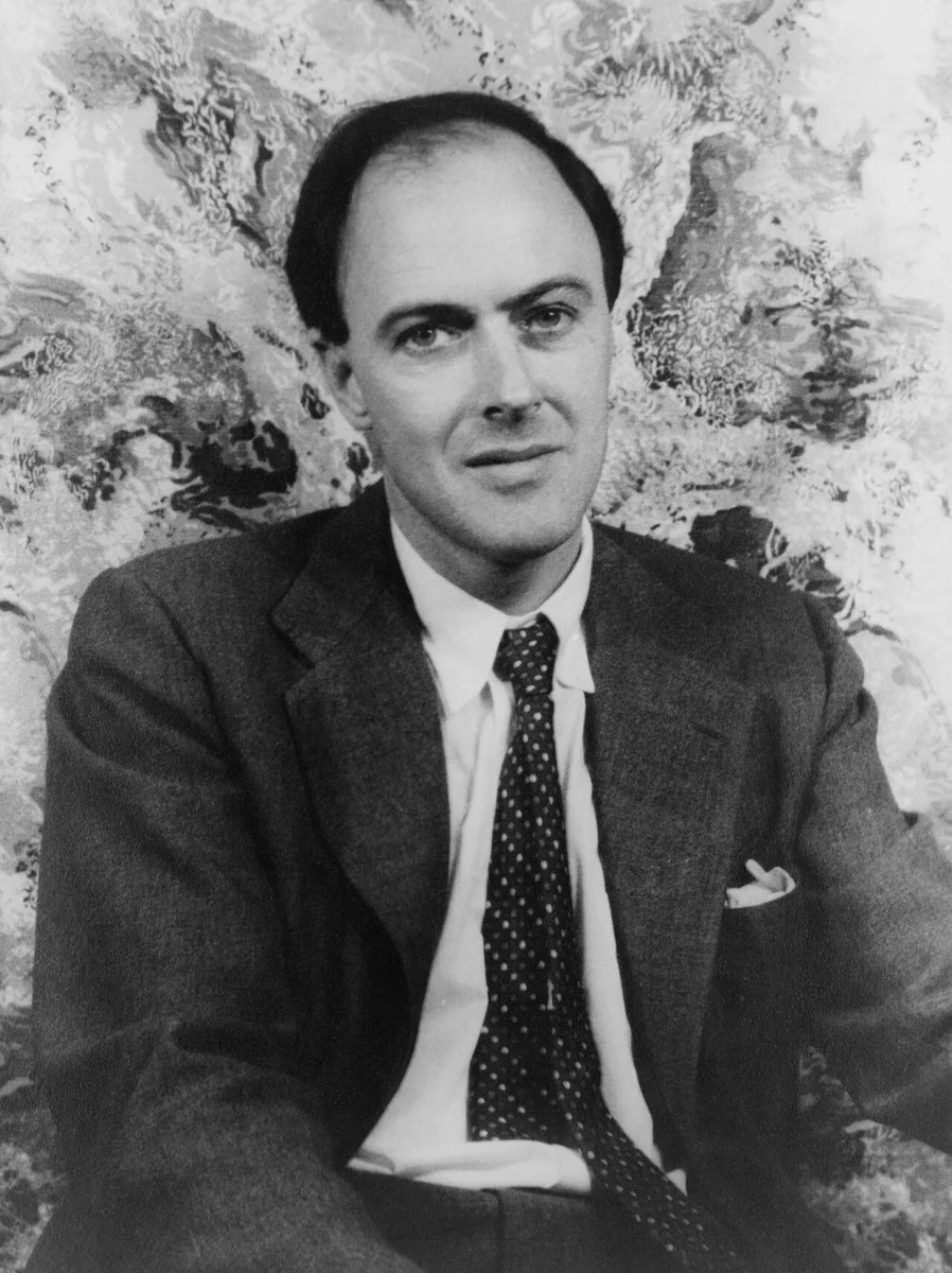 Black and white studio portrait of Roald Dahl in a suit and patterned tie, seated against a mottled backdrop
